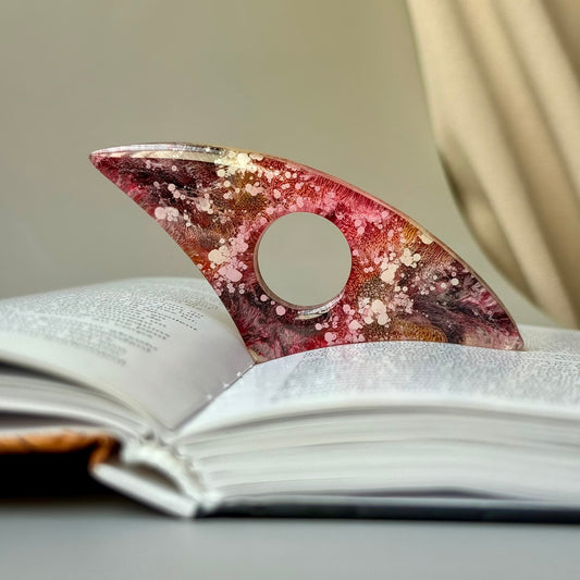 Curved handmade resin page holder in pink and gold alcohol ink, placed in an open book with warm background tones.