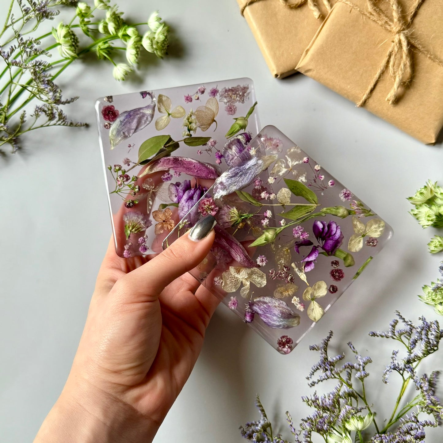 A hand holding two floral resin coasters, showing off the translucent design with purple and ivory pressed flowers.