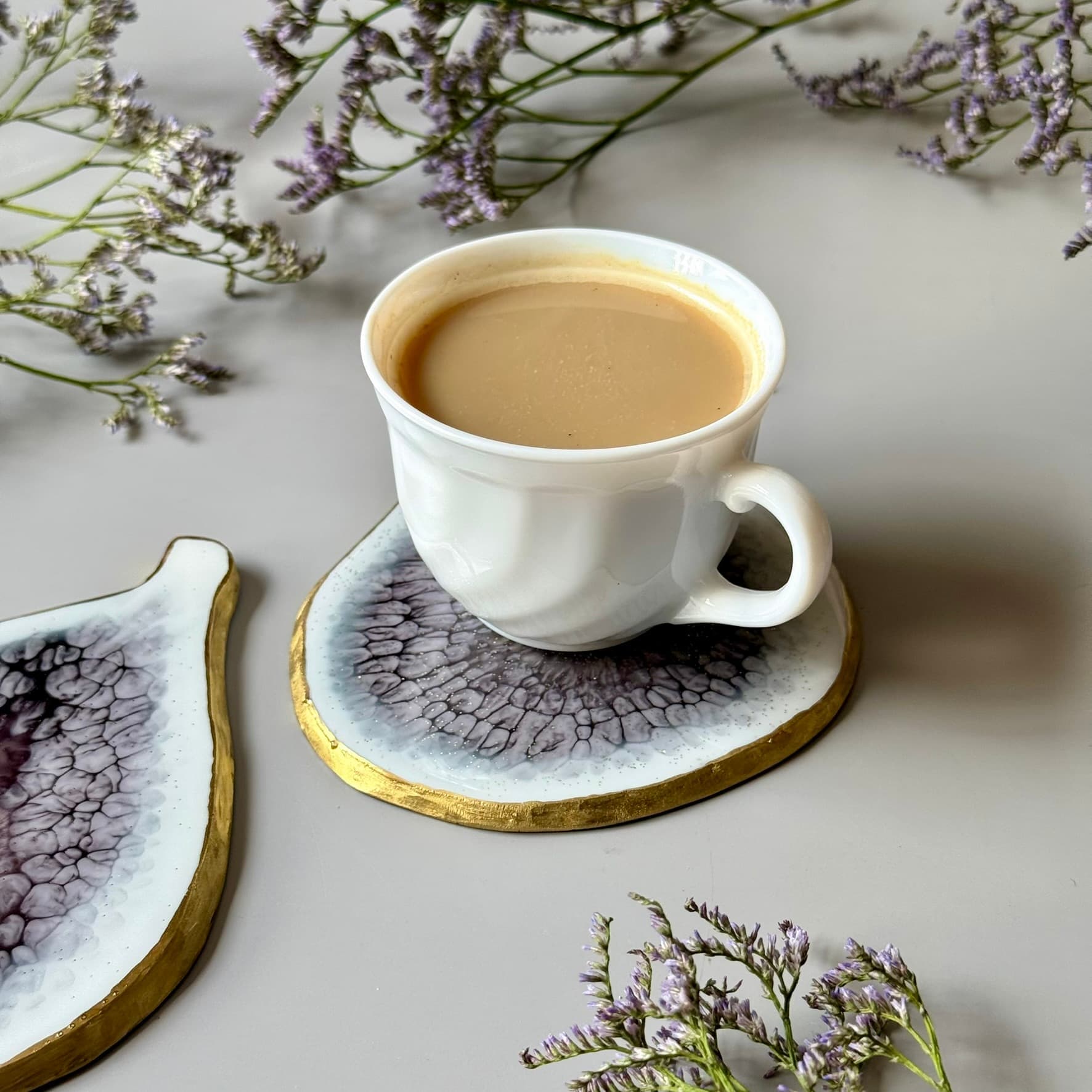 White ceramic cup with a beverage on a decorative coaster, surrounded by purple flowers on a light surface.