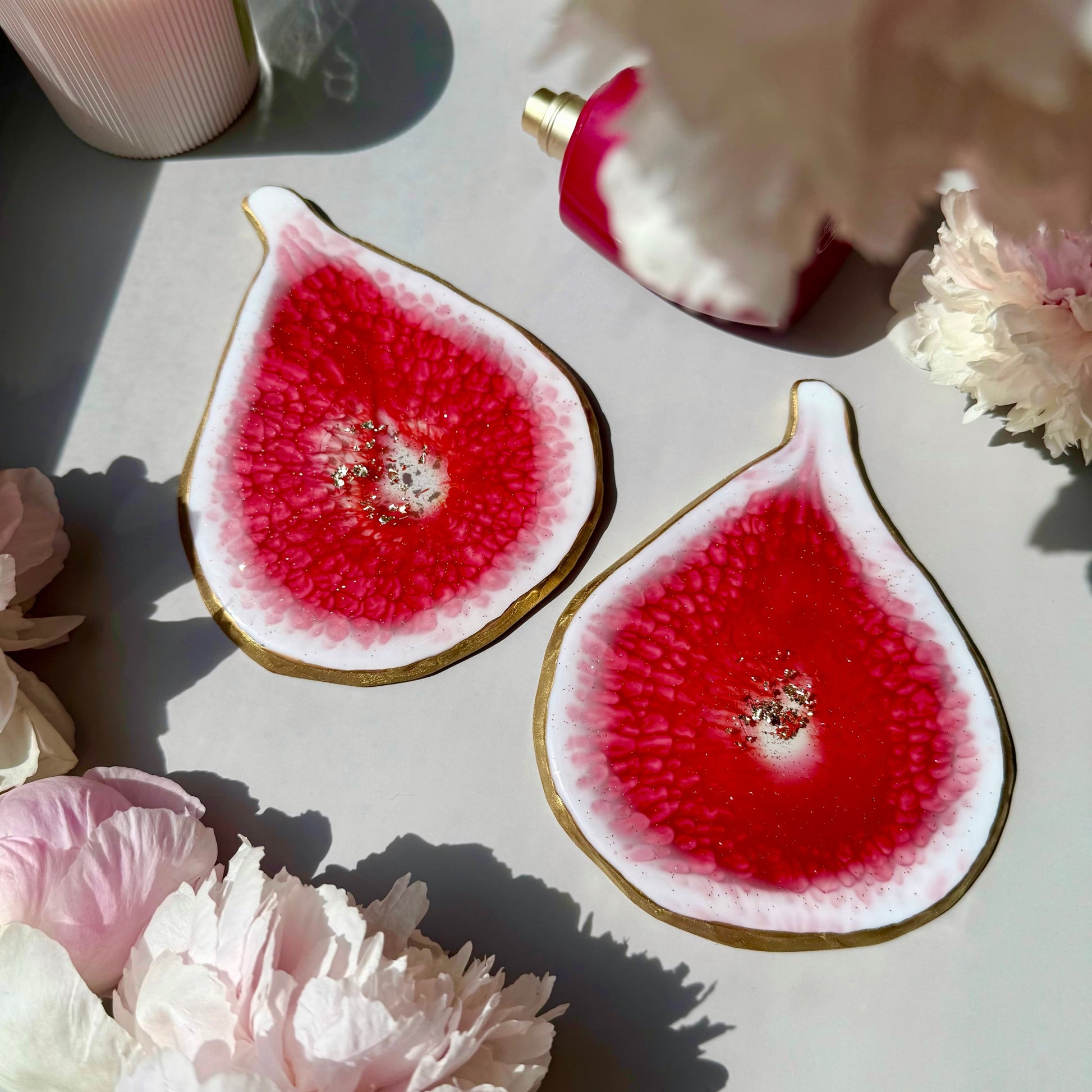 Two handmade fig-shaped coasters with a red interior on a light surface surrounded by flowers.