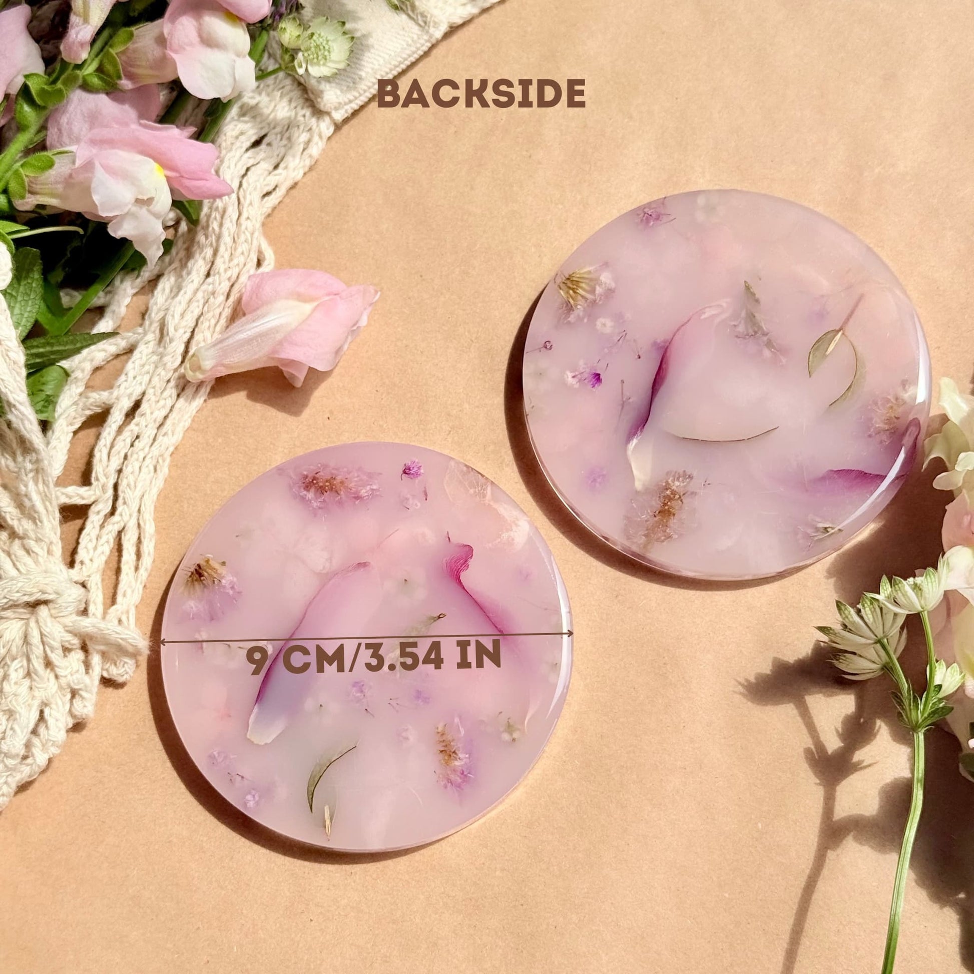 Two round, pink handmade coasters with floral elements on a beige background with flowers around.