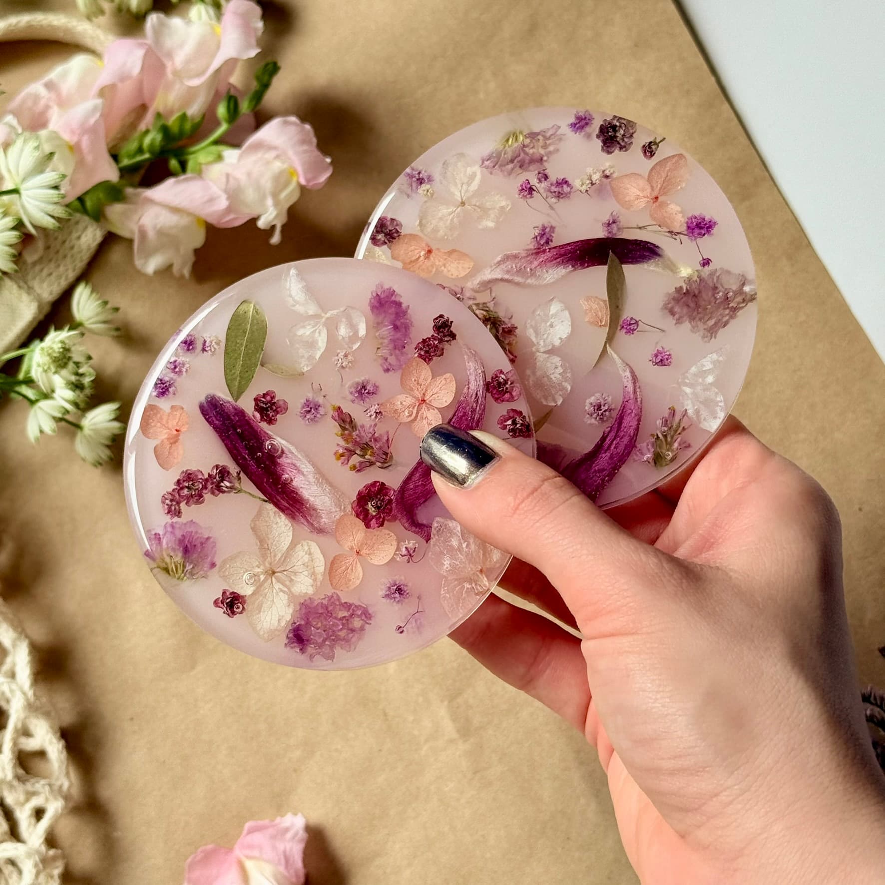 Hand holding a round, pink coasters with floral and leaf embedded, surrounded by flowers on a brown surface.