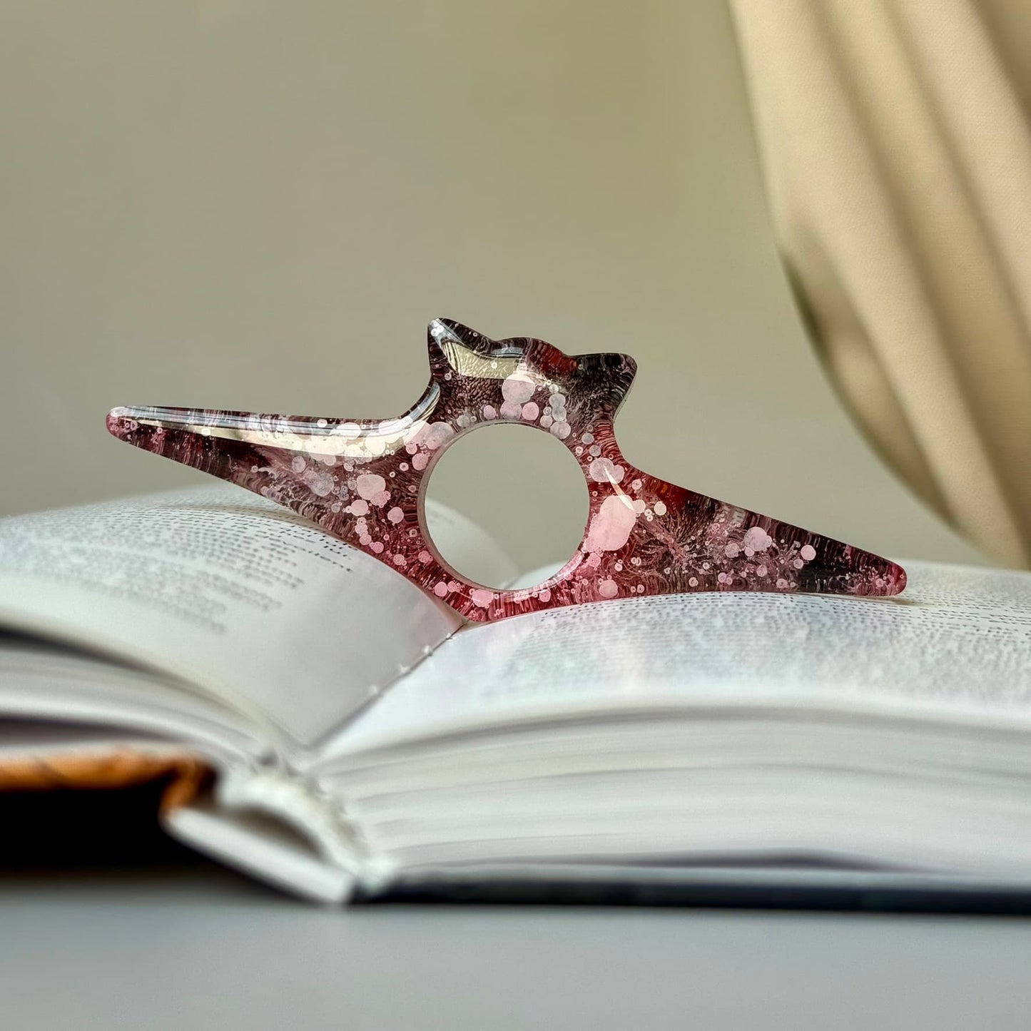 Cat-shaped resin page holder in pink-black ink swirl design, propping open a book for easy reading.