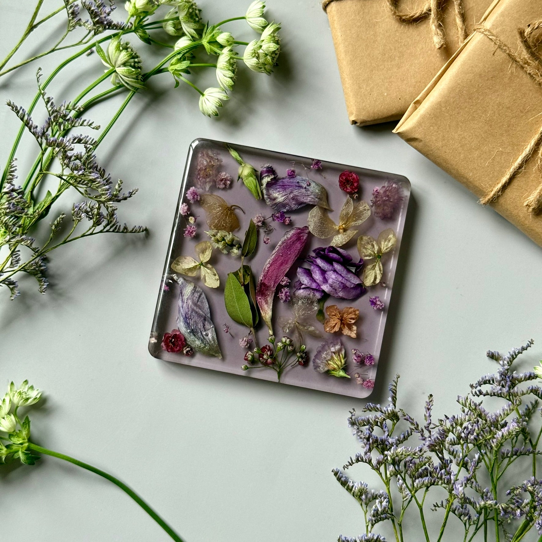 Flat lay of floral resin coaster with vibrant dried flowers on a clean background, surrounded by gift elements and green blooms.