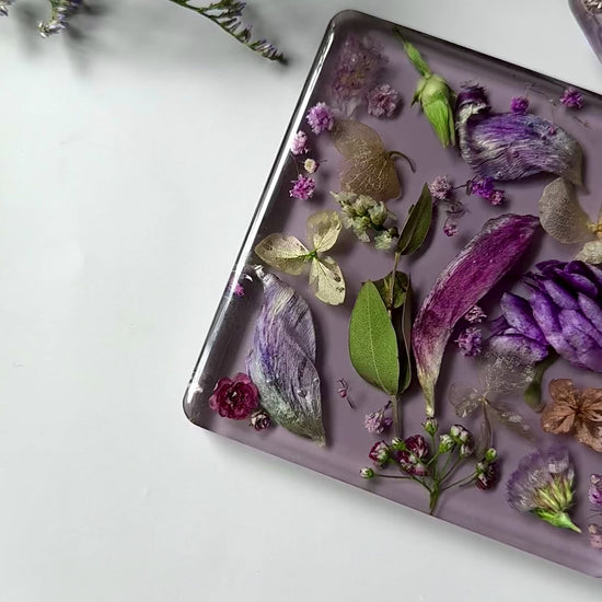 Handmade square resin coasters with real dried flowers in purple and gold tones on a soft lavender background.