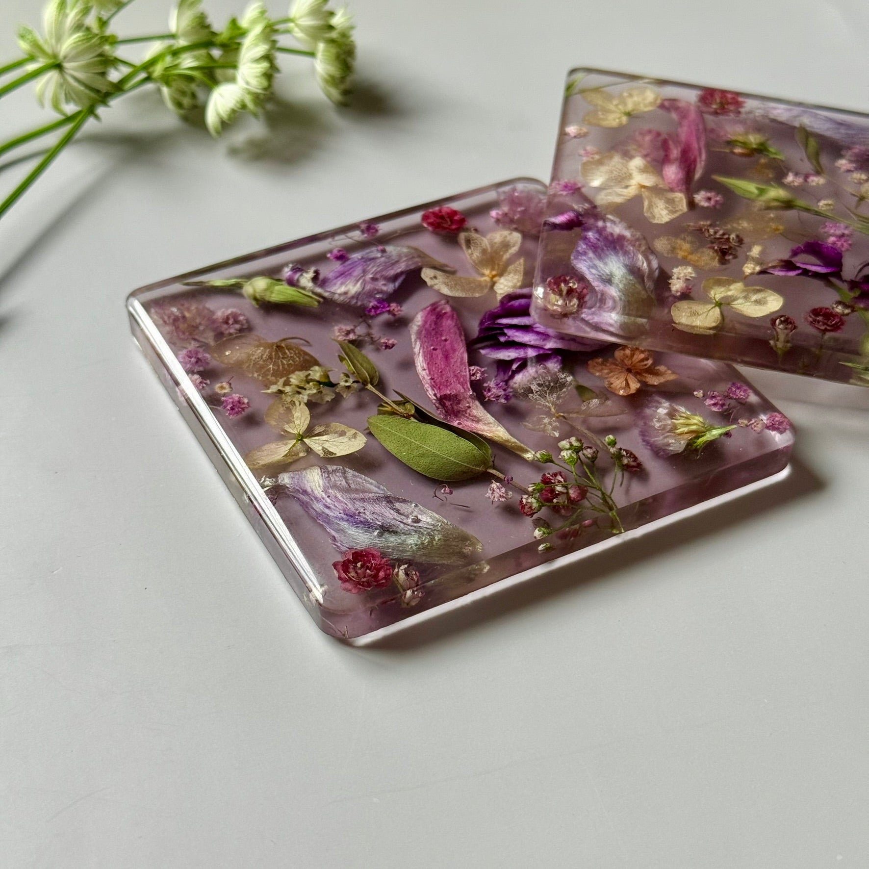 Handmade square resin coasters with real dried flowers in purple and gold tones on a soft lavender background.
